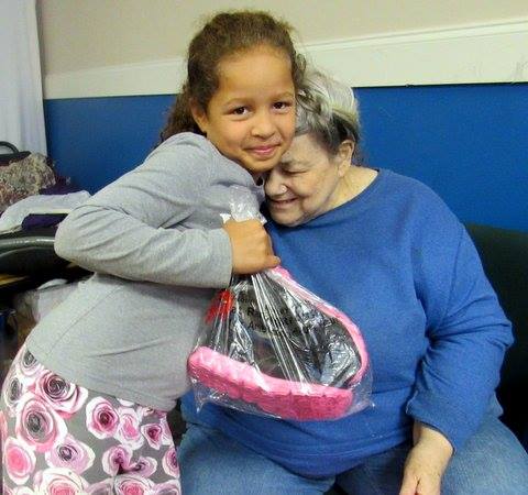 Smiling girl hugs an elderly woman while holding a donated item from Caring Hands Ministries’ community support program.