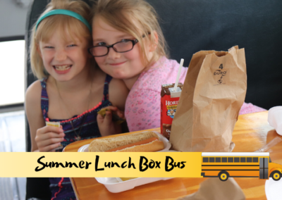 Two young girls smile while sharing a meal in the cafeteria-style seating of the Summer Lunch Box Bus.