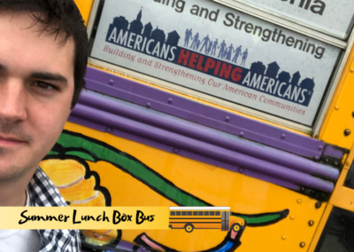 An Of One Accord team member stands by the Americans Helping Americans logo on the Summer Lunch Box Bus.