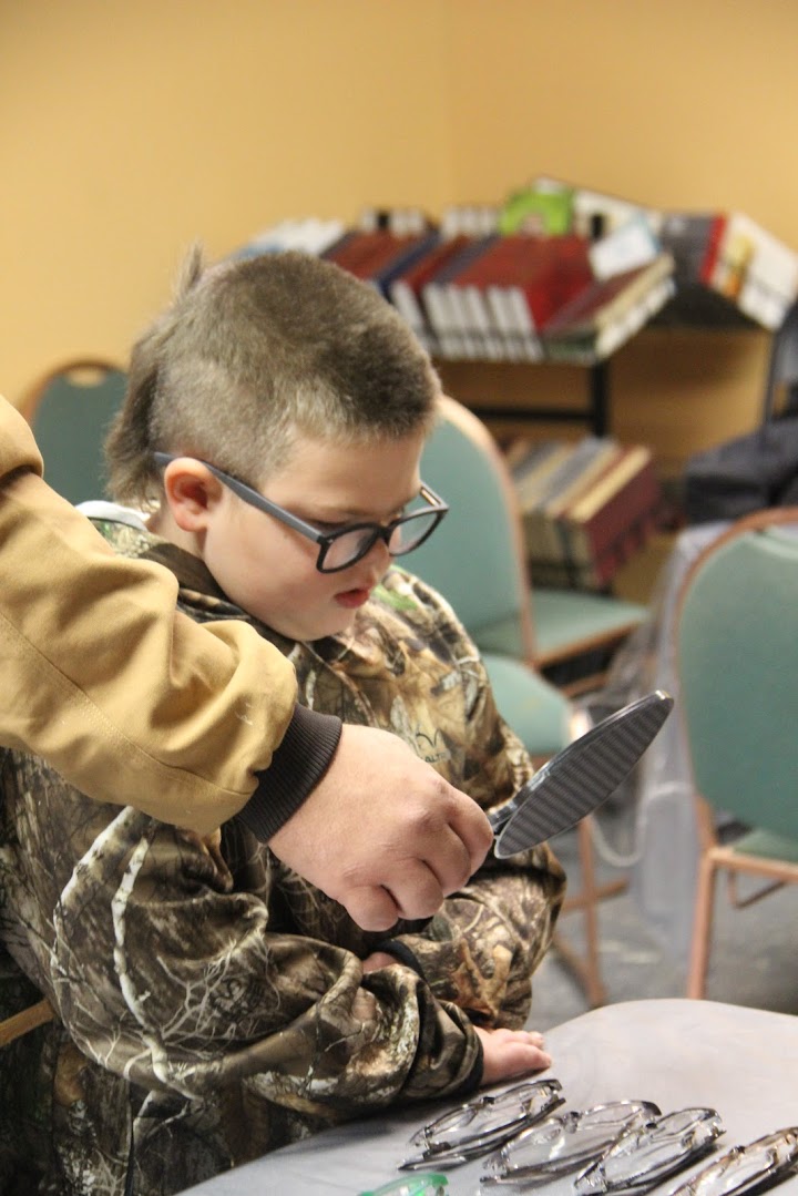 Young boy in camo jacket trying on new prescription glasses at Of One Accord free eyeglass clinic.