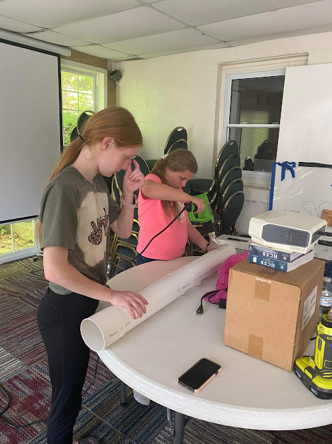 Two girls use a power drill to cut holes in a long PVC pipe for a school hydroponic garden project