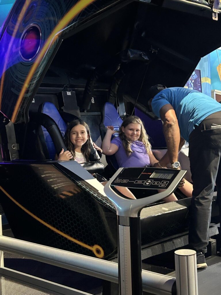 Two girls smiling in a motion simulator ride at an entertainment center during an Americans Helping Americans summer camp.