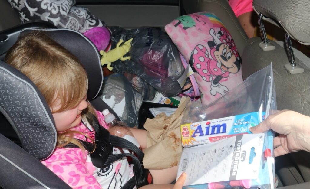 Young girl in car seat receiving Americans Helping Americans Mighty Molars dental kit for better oral health.