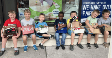 A group of happy young boys sits on a storefront step, holding boxes and showing off their brand new shoes.